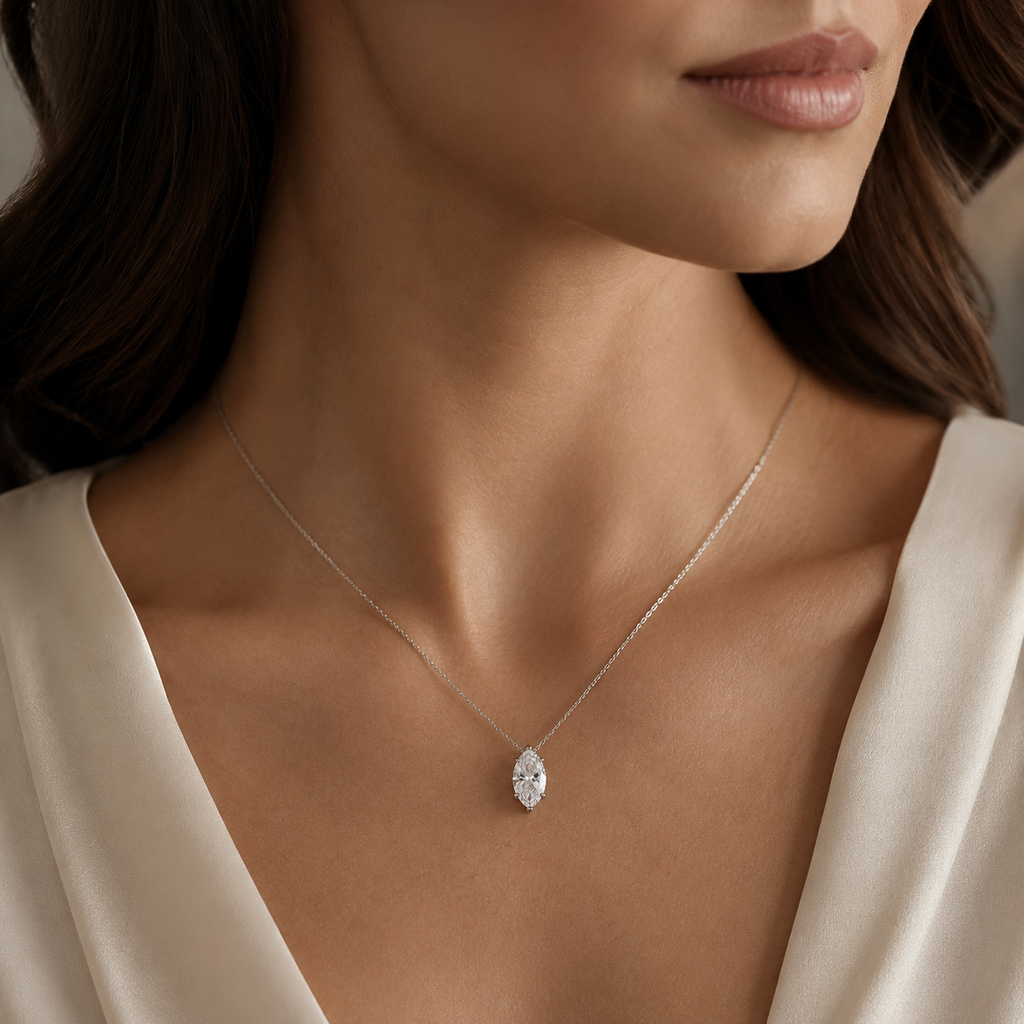 A close-up of a woman wearing a delicate silver chain necklace with a marquise-cut clear gemstone pendant, styled with a cream satin top.